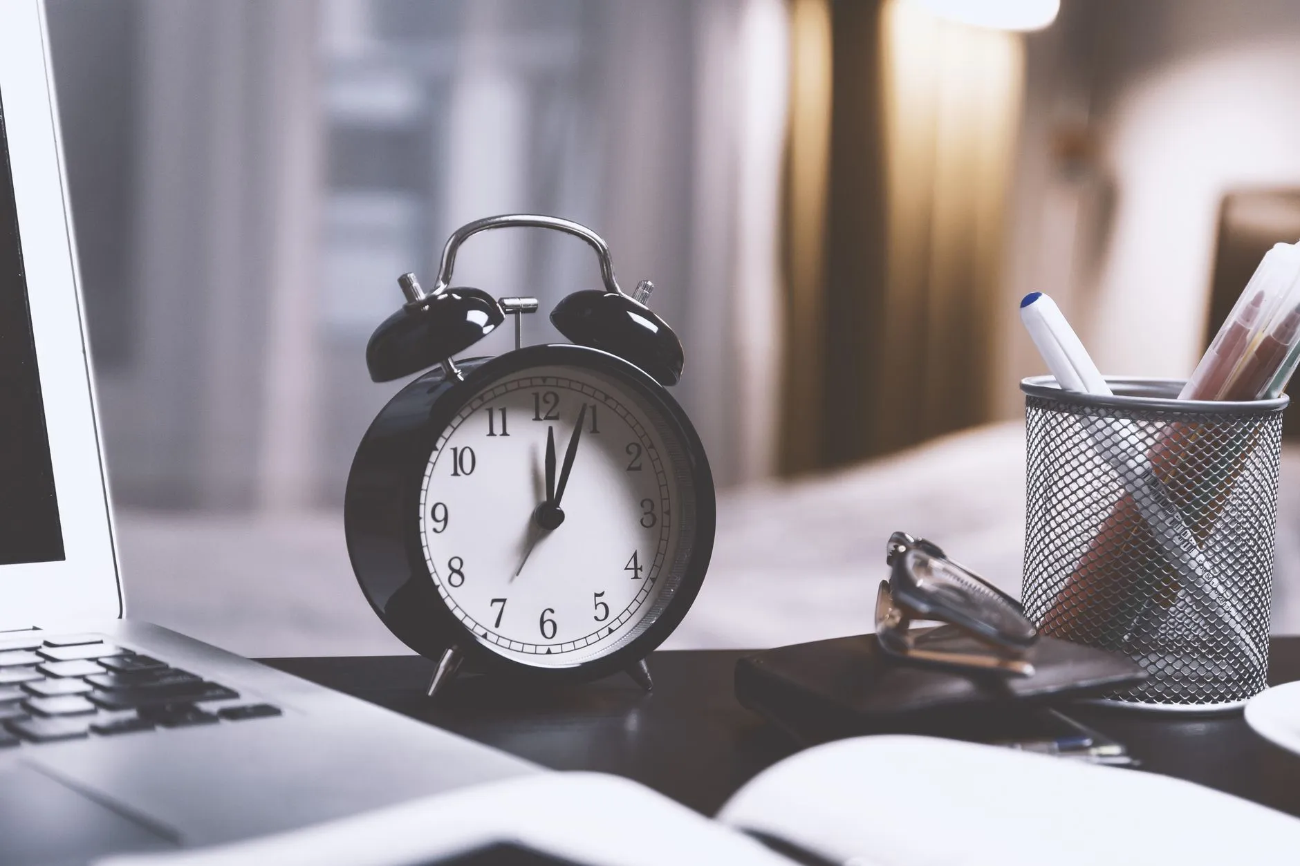 A classic analog alarm clock on a modern desk setup with a laptop and pens.